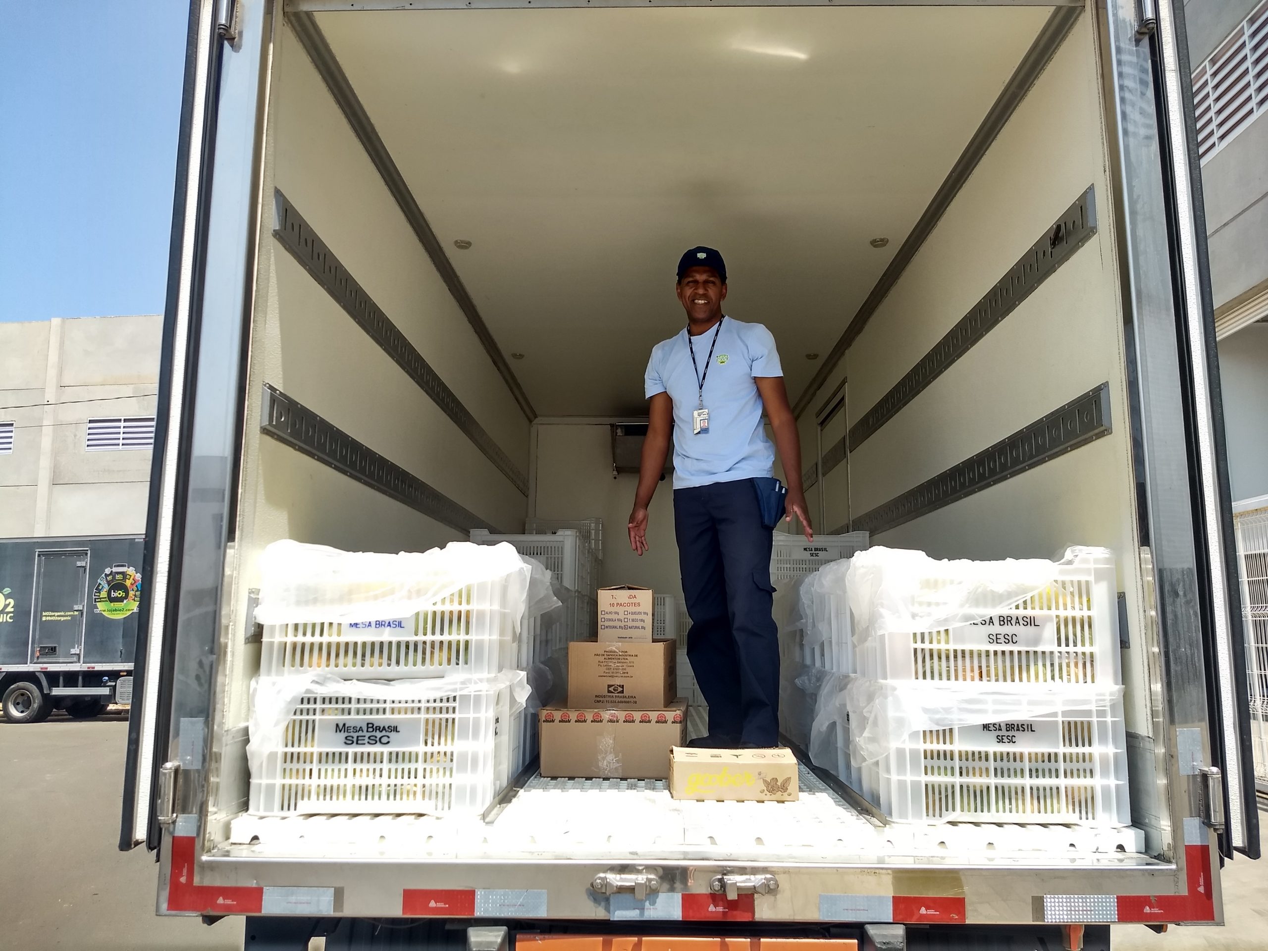 Funcionário do Mesa Brasil recebendo doações da Lourenço, ele está dentro do Baú do Caminhão do Mesa Brasil, sorrindo, com as caixas de doação da Lourenço Alimentos no chão.